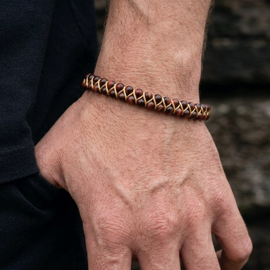 Close-up of a man's wrist wearing a braided red tigers eye bracelet with a blurred natural background.