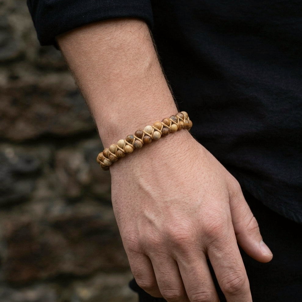 A man's hand wearing a beaded picture jasper bracelet with a stone wall background