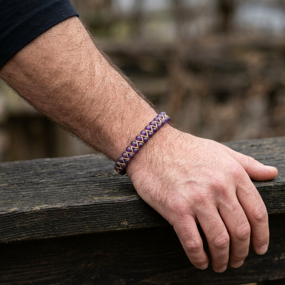 A man's hand wearing a braided purple amethyst bracelet on a wooden surface with a blurred natural background