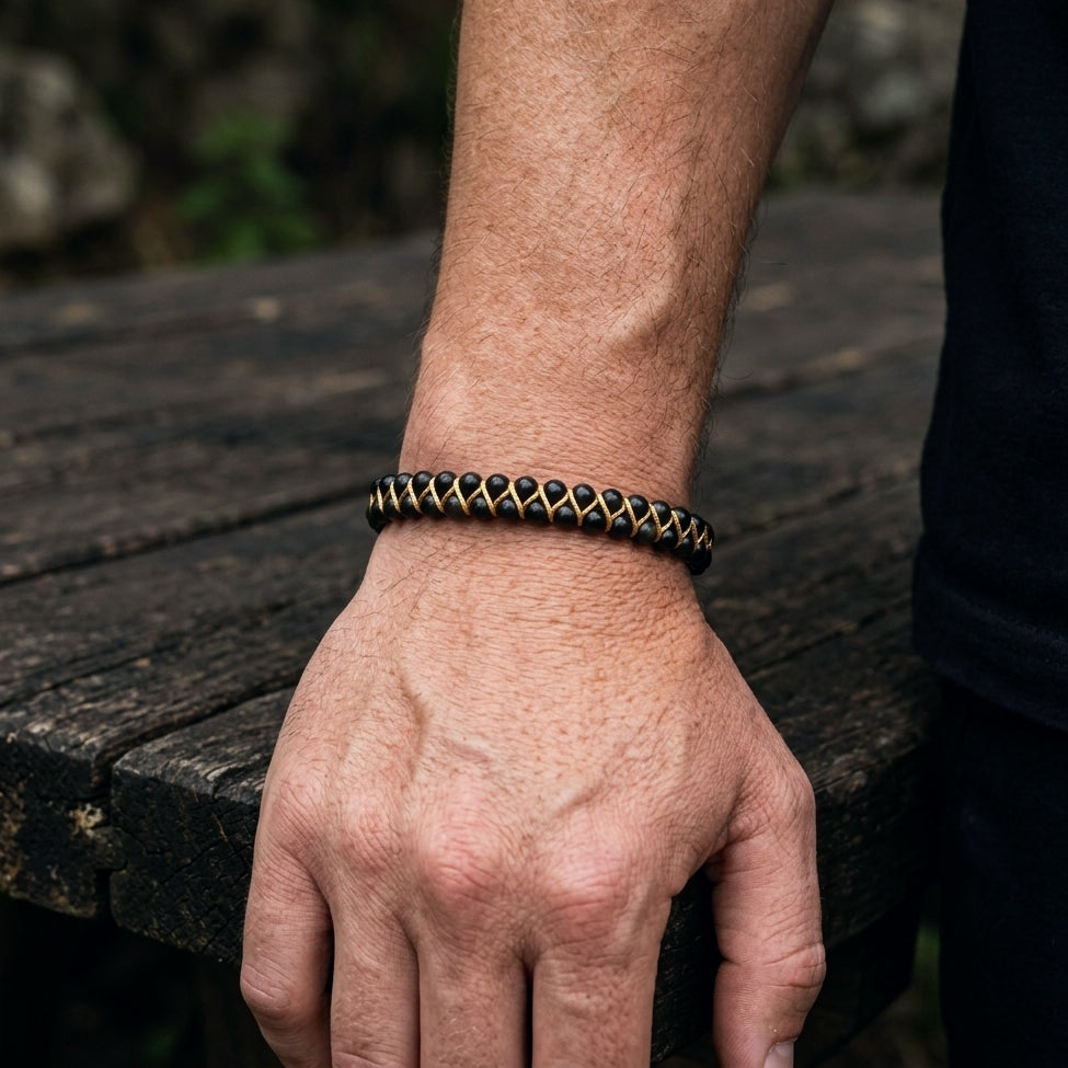 A man's hand wearing a black obsidian and gold bracelet on a wooden surface with a blurred natural background
