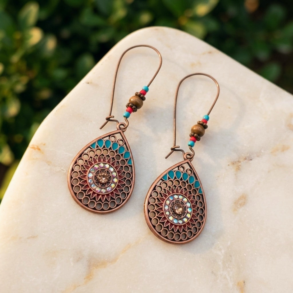 A pair of bohemian-style teardrop dangle earrings with intricate filigree detailing, turquoise and red accents, and small bead strands on copper-toned hooks, displayed on a light marble surface with a blurred green background.