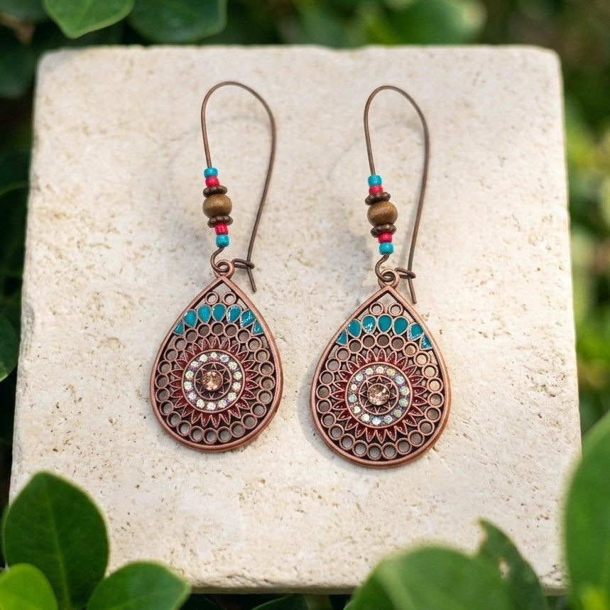 A pair of bohemian-style copper teardrop dangle earrings with mandala flowers and turquoise and red bead accents, displayed on a light stone slab surrounded by green foliage.