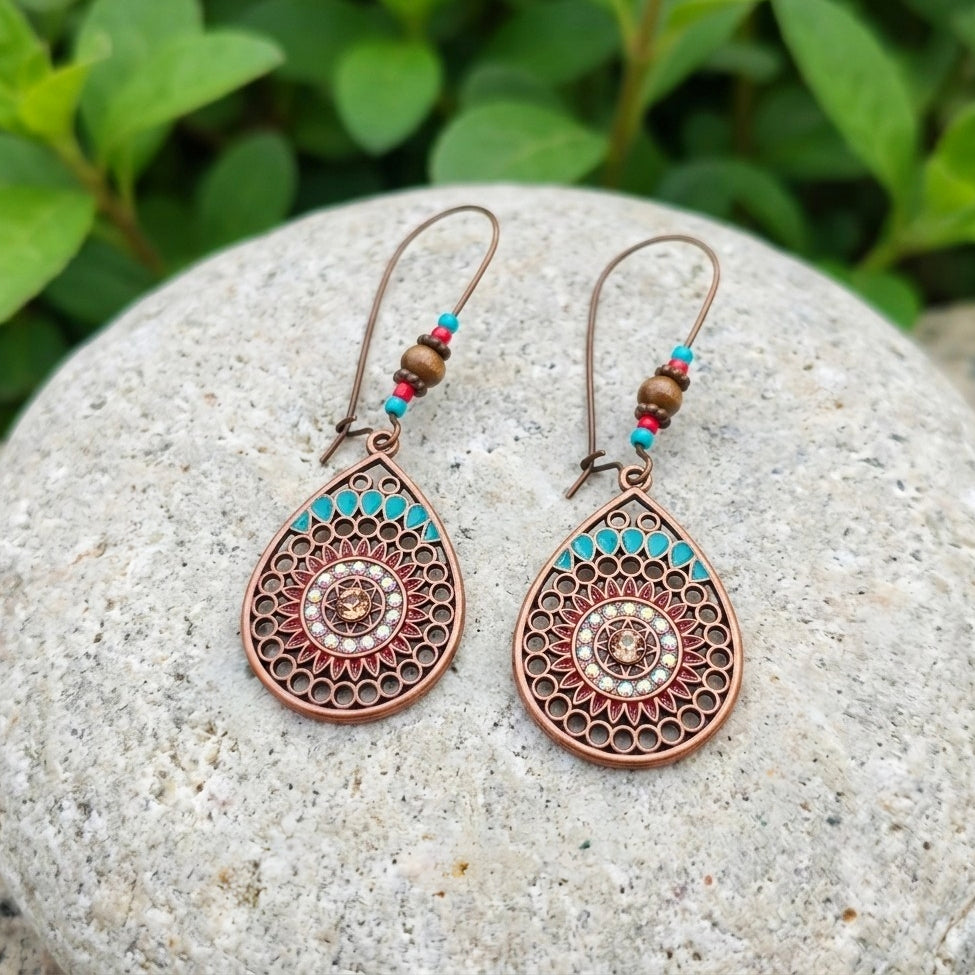 A pair of bohemian-style copper teardrop dangle earrings with a mandala flower and turquoise and red bead accents, displayed on a round gray stone with green foliage in the background.