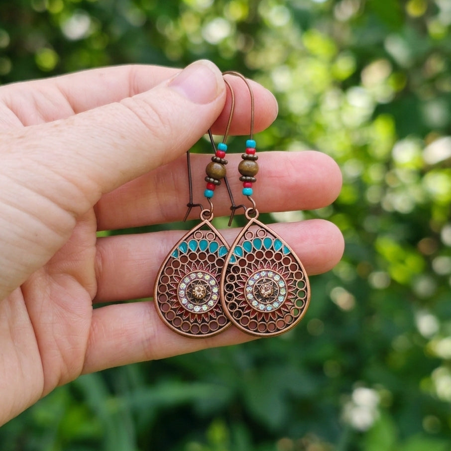 A pair of copper-toned teardrop dangle earrings with turquoise and brown bead details, held in a hand against a blurred green outdoor background.