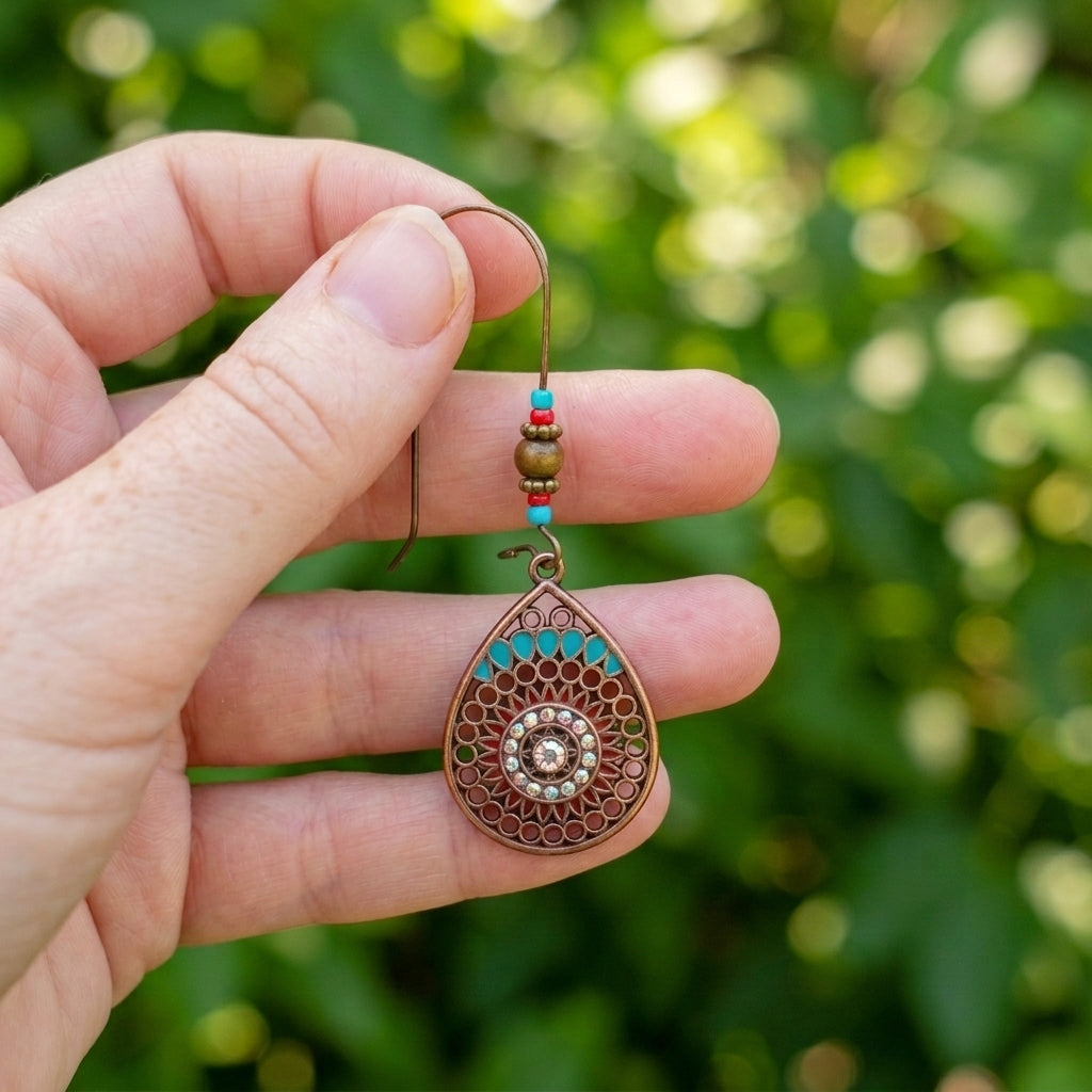 A hand holding a teardrop-shaped bohemian-style dangle earring with a bronze filigree pattern, turquoise accents, and small colorful beads, set against a blurred green outdoor background.