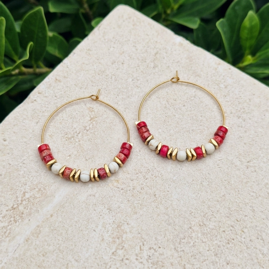 Gold hoop earrings with red sea sediment jasper and white beads, displayed on a light stone surface with green foliage in the background. 