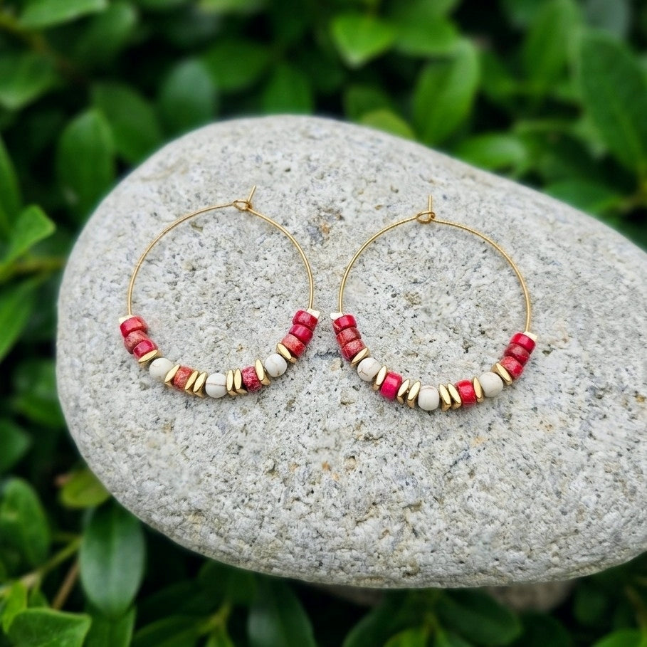 Gold hoop earrings with red jasper and cream beads, displayed on a gray stone with green foliage in the background.