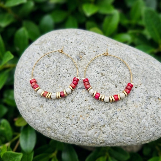 Gold hoop earrings with red jasper and cream beads, displayed on a gray stone with green foliage in the background.