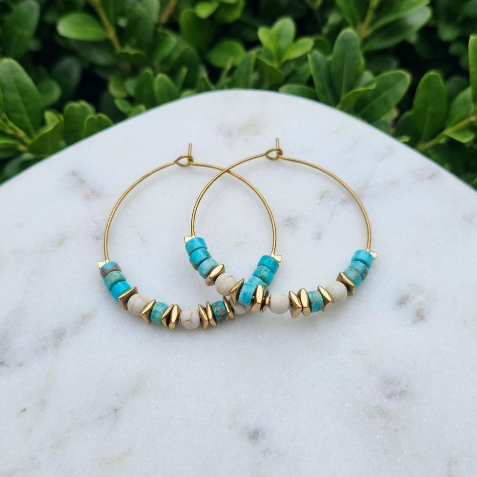 Gold hoop earrings with turquoise sea sediment jasper and white beads, displayed on a white marble surface with green foliage in the background.
