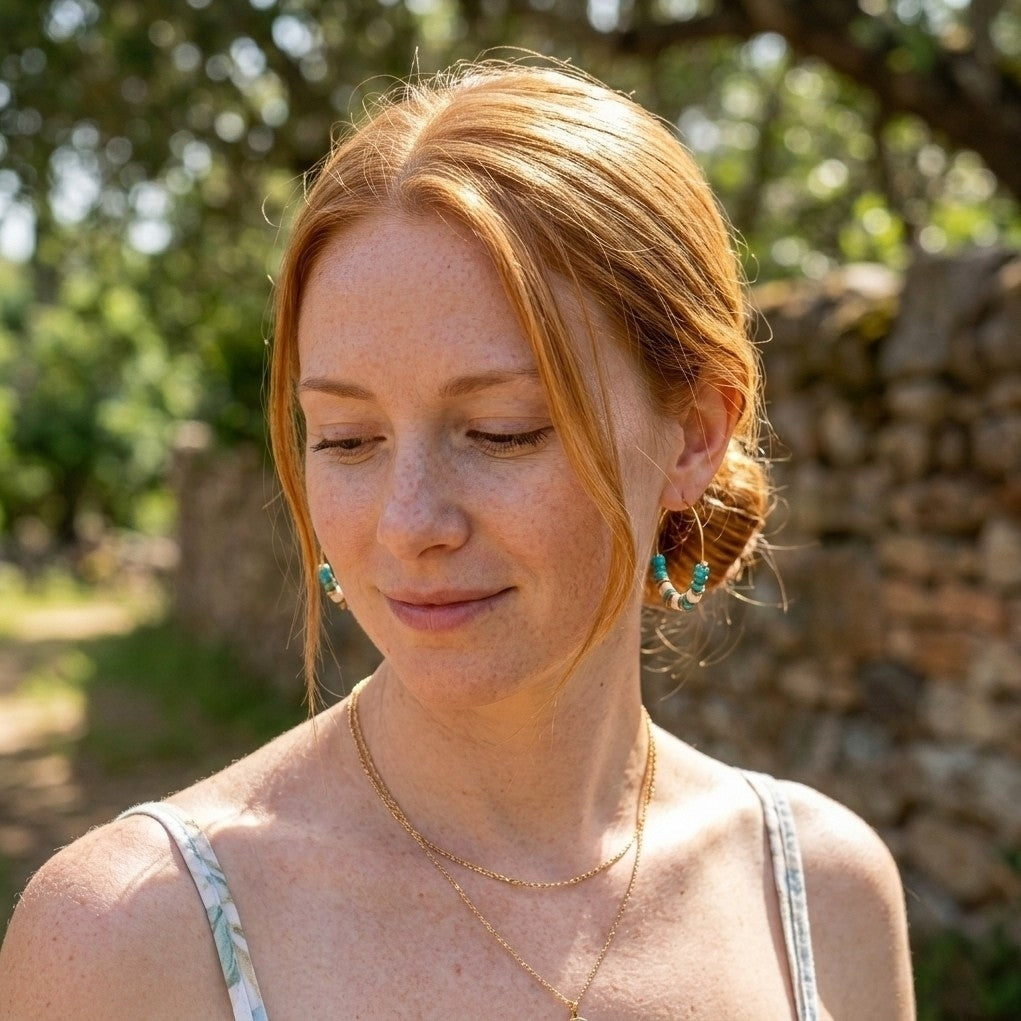 A woman with red hair looking down, wearing a white floral spaghetti strap top, layered gold necklaces, and turquoise drop earrings, set against a blurred outdoor background of trees and a stone wall.