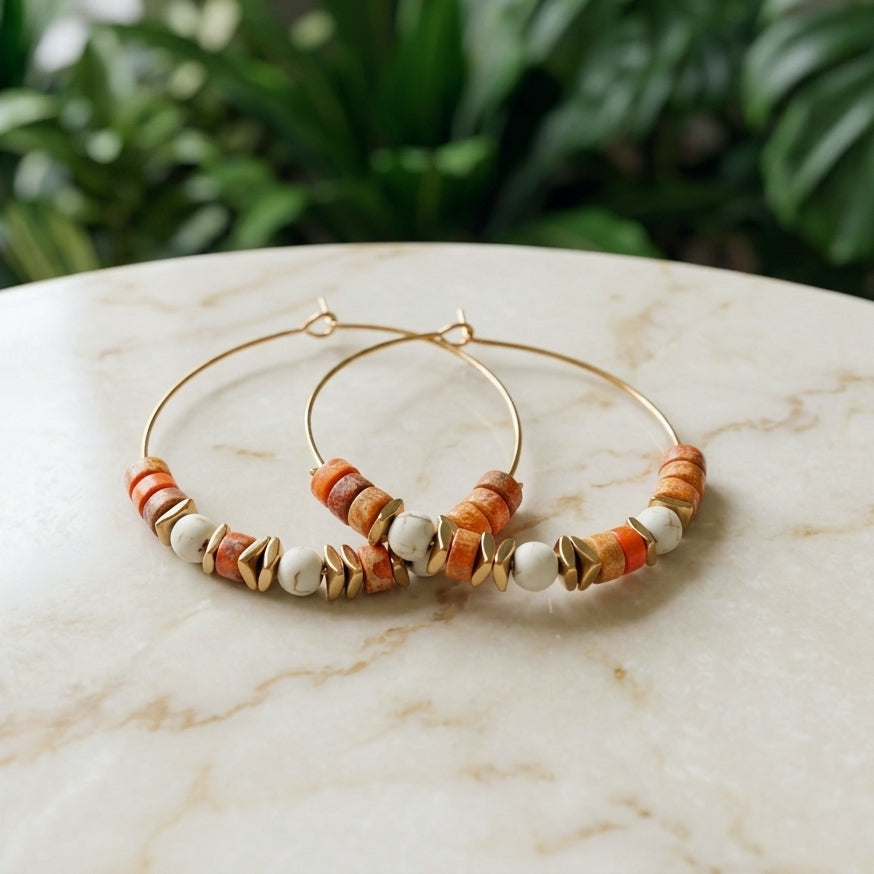 A pair of gold hoop earrings with orange and white beaded details, displayed on a light marble surface with a blurred green plant background.