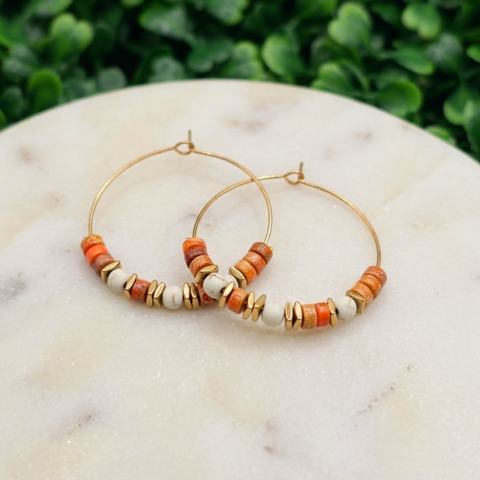 Gold hoop earrings with orange and white beads, displayed on a white marble surface with a blurred green foliage background.