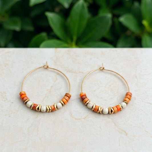 A pair of gold hoop earrings with small orange sea sediment jasper, white, and brown beads, displayed on a light marble surface with a blurred green plant background.