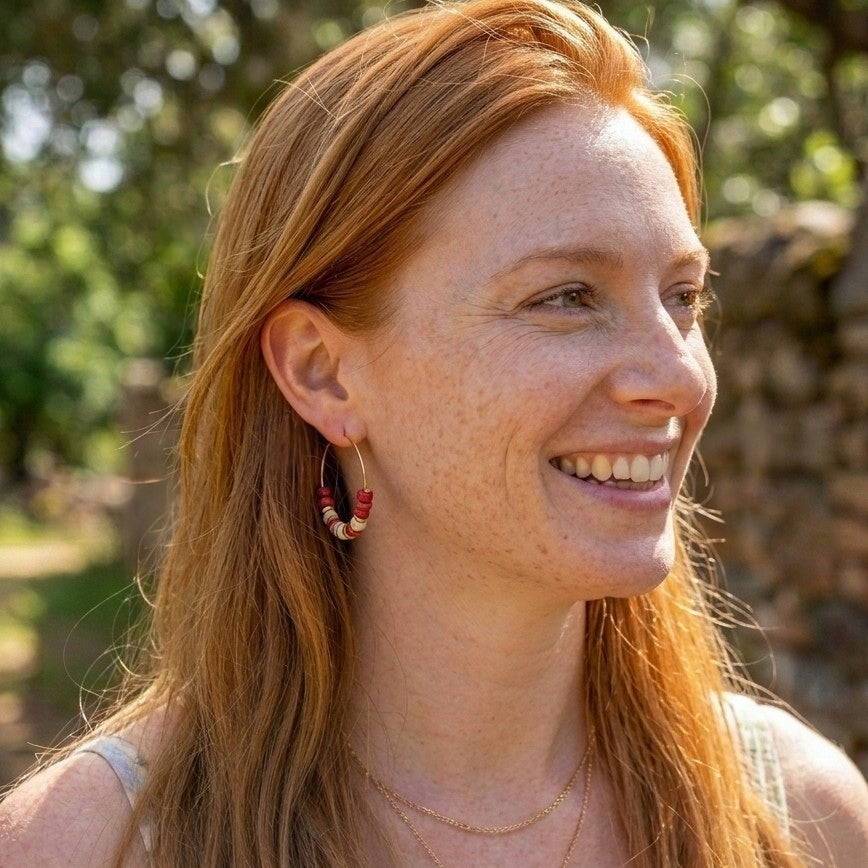 A smiling woman with long red hair wearing a small gold hoop earring with red and white bead details, photographed outdoors with a blurred green and stone background.