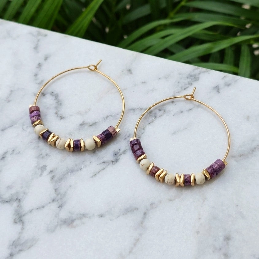 Gold hoop earrings with purple and cream beads, displayed on a white marble surface with green foliage in the background.