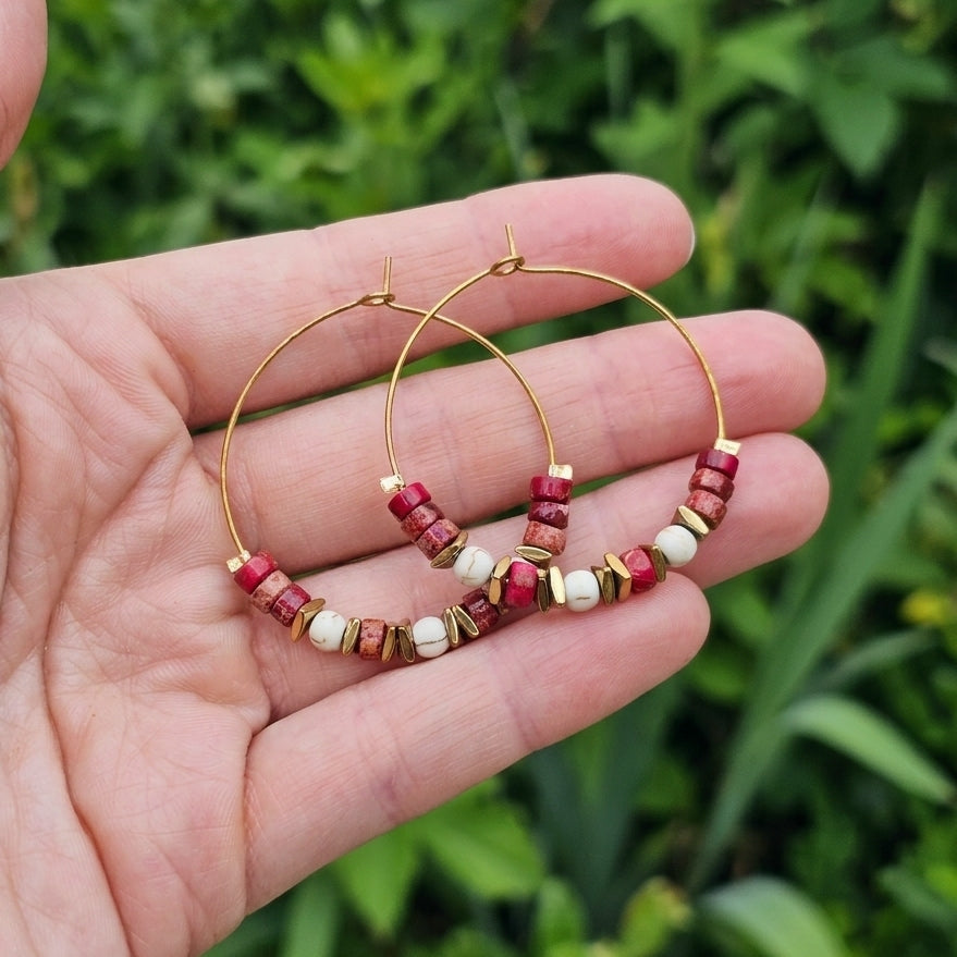 Gold hoop earrings with red jasper gemstone and white beaded details, held in a person's hand against a blurred green outdoor background.