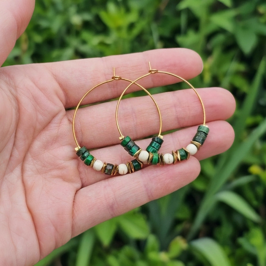 Gold hoop earrings with green jasper gemstone and white beaded accents, held in a person's hand against a blurred green outdoor background.