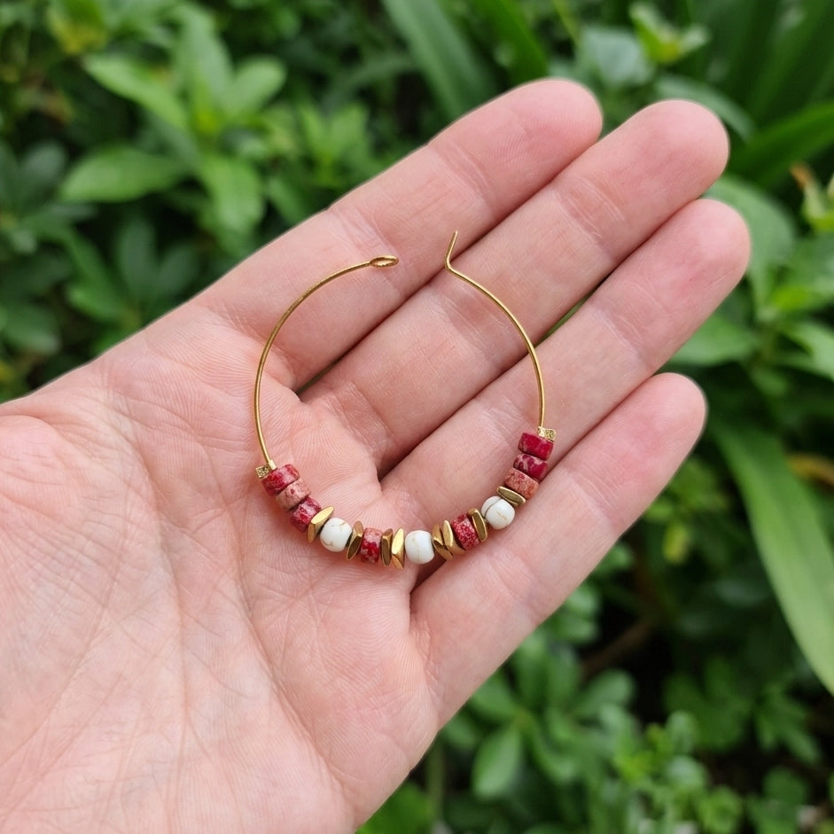A delicate gold hoop earring with small red and white beads, held in a person's hand against a blurred green outdoor background.
