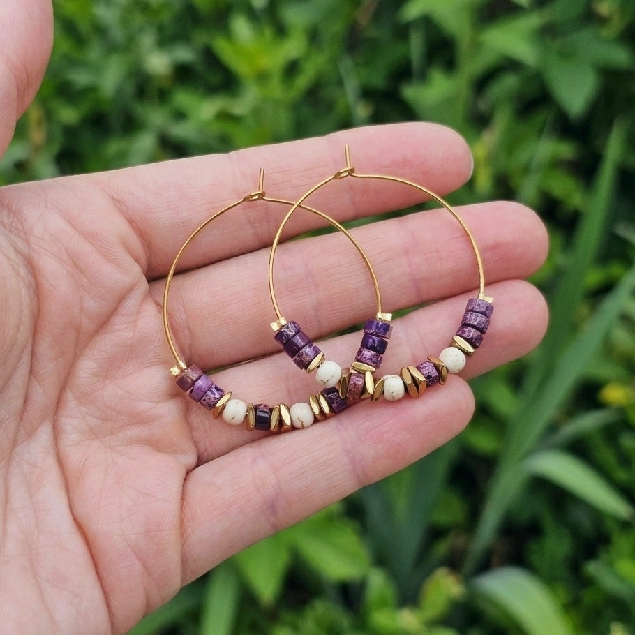 Gold hoop earrings with purple jasper and white beads, held in a person's hand against a blurred green outdoor background.