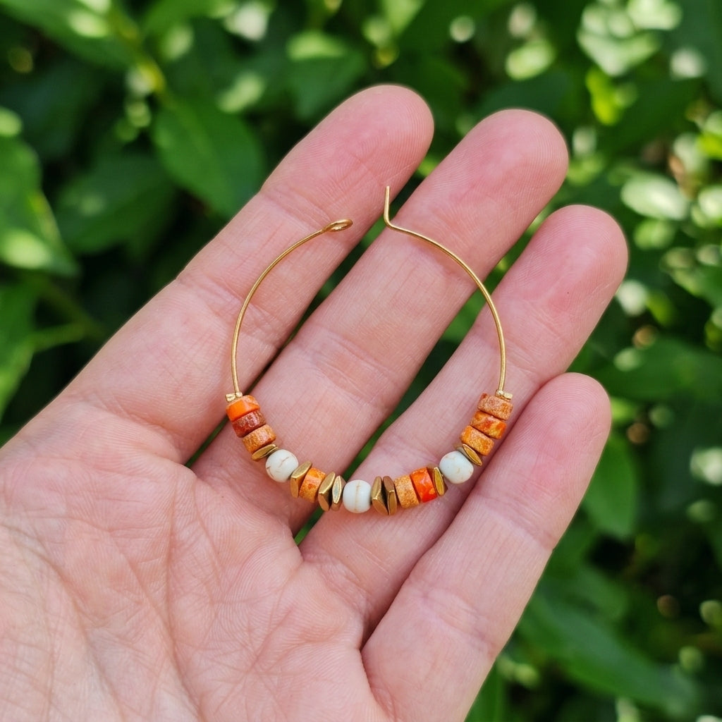 A small gold hoop earring with a row of orange, brown, and white wooden beads, held in a person's hand against a blurred green leaf background.