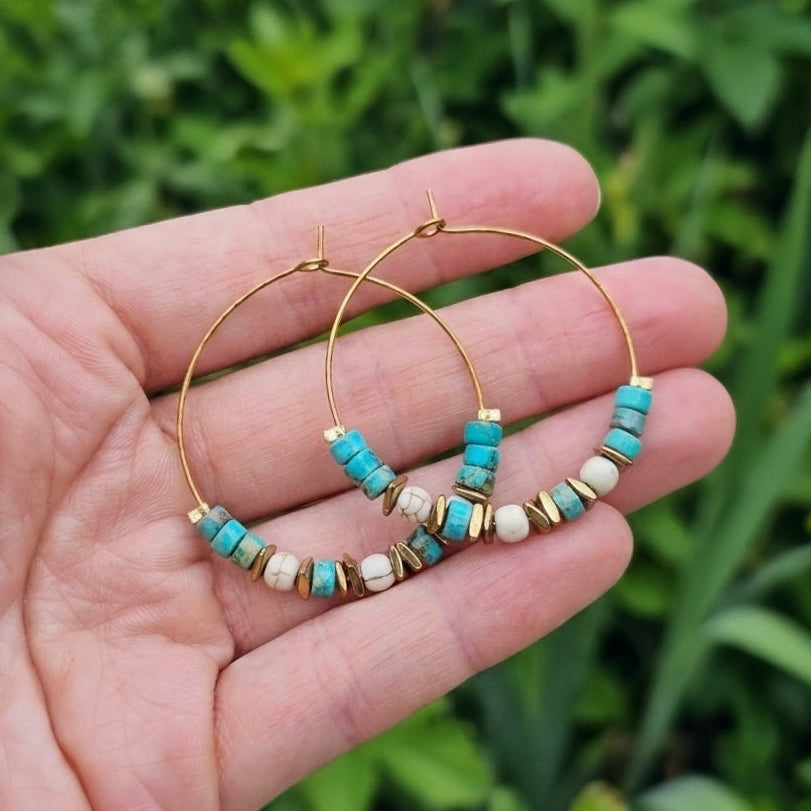 Gold hoop earrings with turquoise jasper and white beads, held in a person's hand against a blurred green outdoor background.