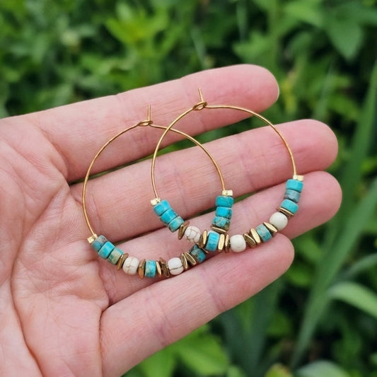 Gold hoop earrings with turquoise jasper and white beads, held in a person's hand against a blurred green outdoor background.