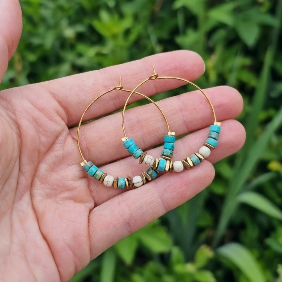 Gold hoop earrings with turquoise jasper and white beads, held in a person's hand against a blurred green outdoor background.