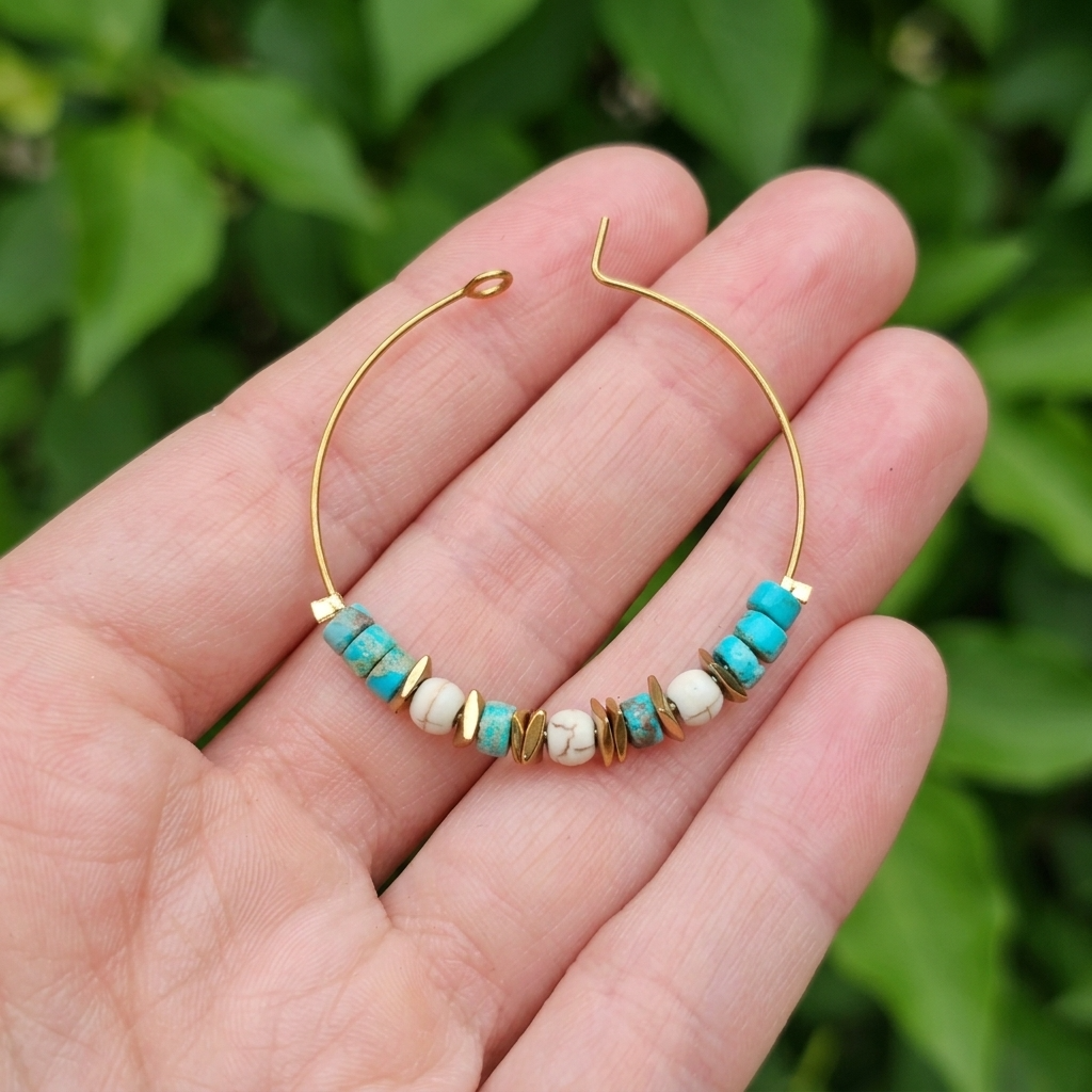 A gold hoop earring with turquoise and white beads, held in a person's hand against a blurred green leaf background.