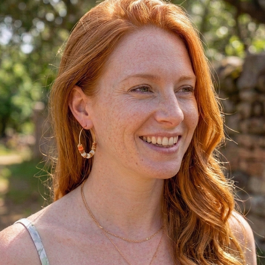 A smiling woman with long wavy red hair wearing gold hoop earrings with orange and white beads, layered gold necklaces, and a light-colored spaghetti strap top, photographed outdoors with a blurred green and stone background.