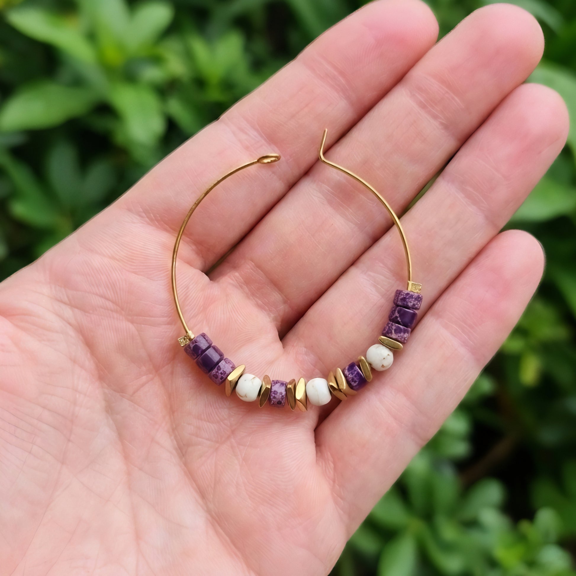 A gold wire hoop earring with purple jasper and white beads, held in a person's hand against a blurred green outdoor background.
