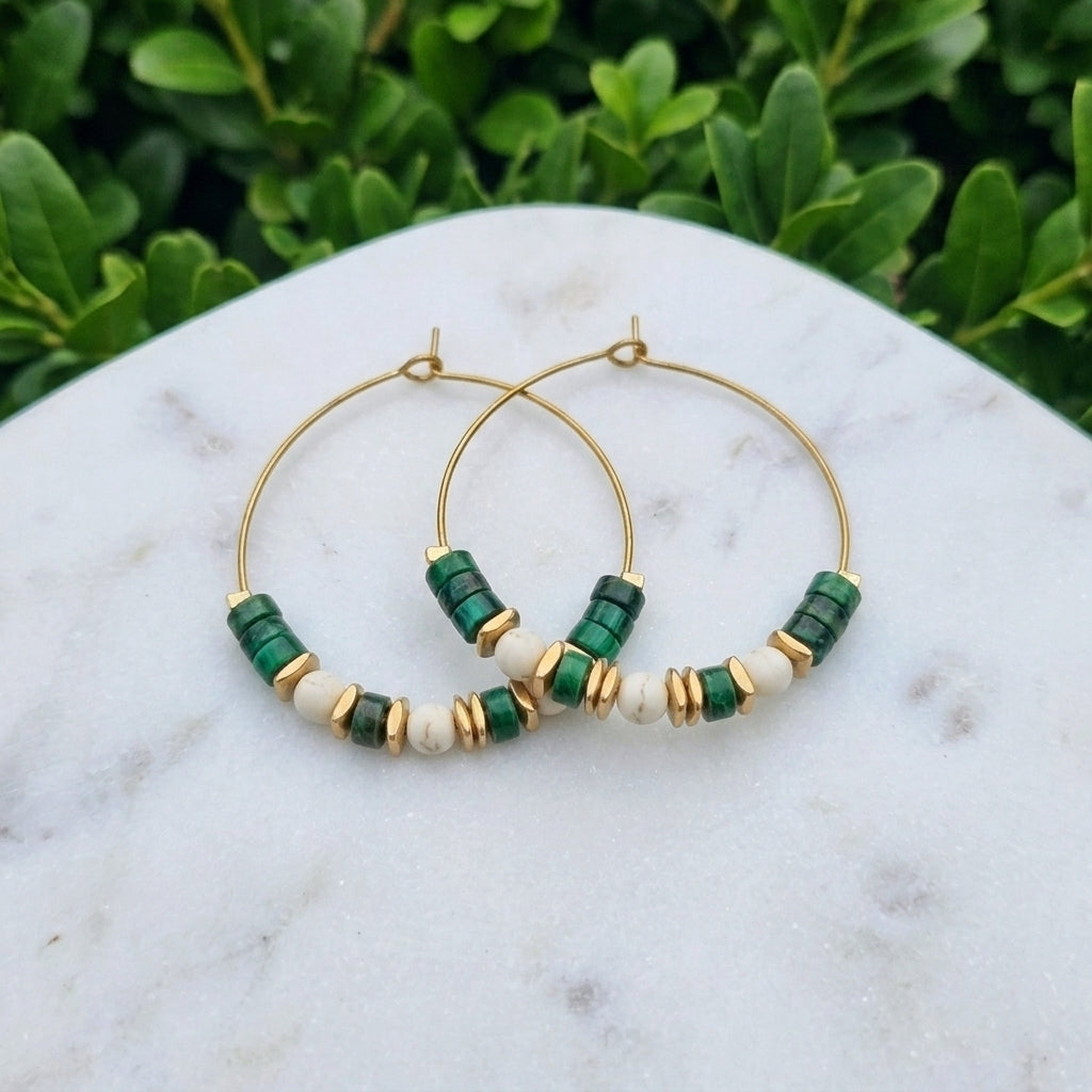 Gold hoop earrings with green and white beads, displayed on a white marble surface with green foliage in the background.