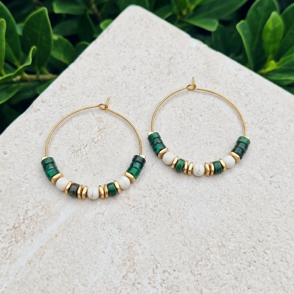 A pair of gold hoop earrings with green  jasper and white beads, displayed on a light stone surface with green foliage in the background.