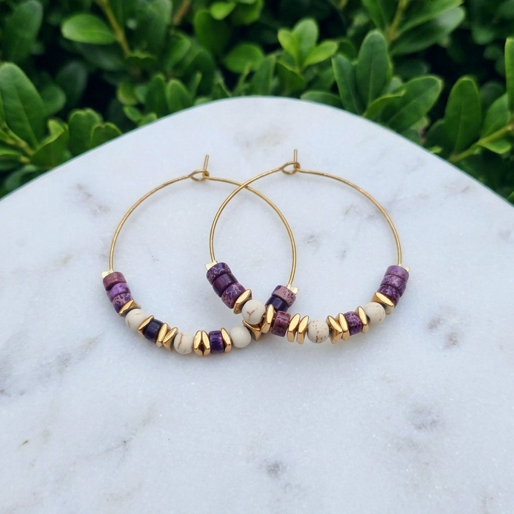 Gold hoop earrings with purple and cream beads, displayed on a white marble surface with green foliage in the background.