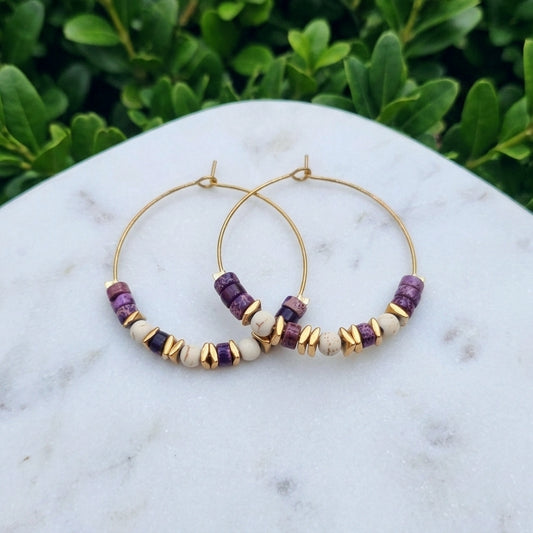 Gold hoop earrings with purple and cream beads, displayed on a white marble surface with green foliage in the background.