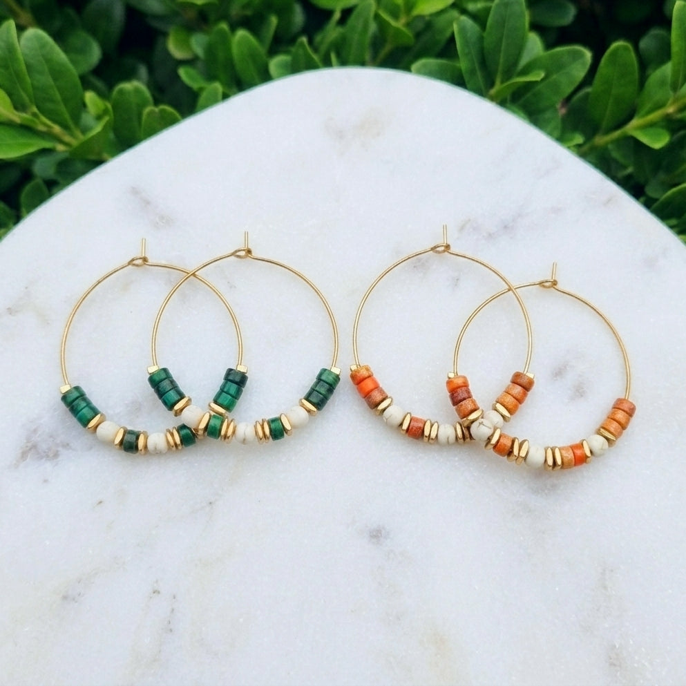 Two pairs of gold hoop earrings with green and orange jasper bead accents, displayed on a white marble surface with green foliage in the background.