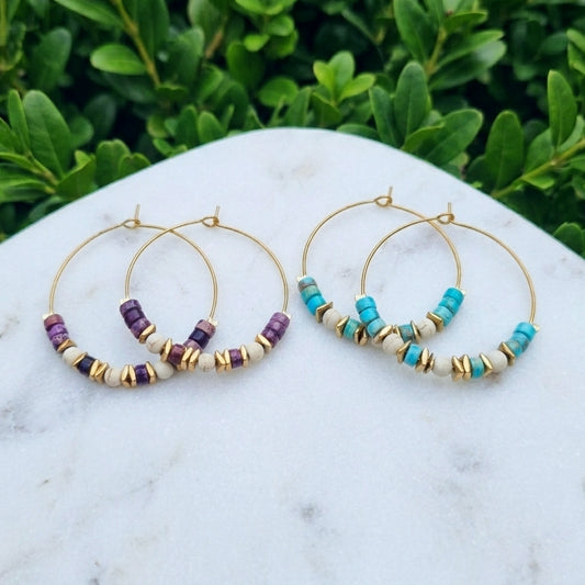 Two pairs of gold hoop earrings with small gold spacer beads and clusters of purple and turquoise sea sediment jasper and white beads, displayed on a white marble surface with green foliage in the background.