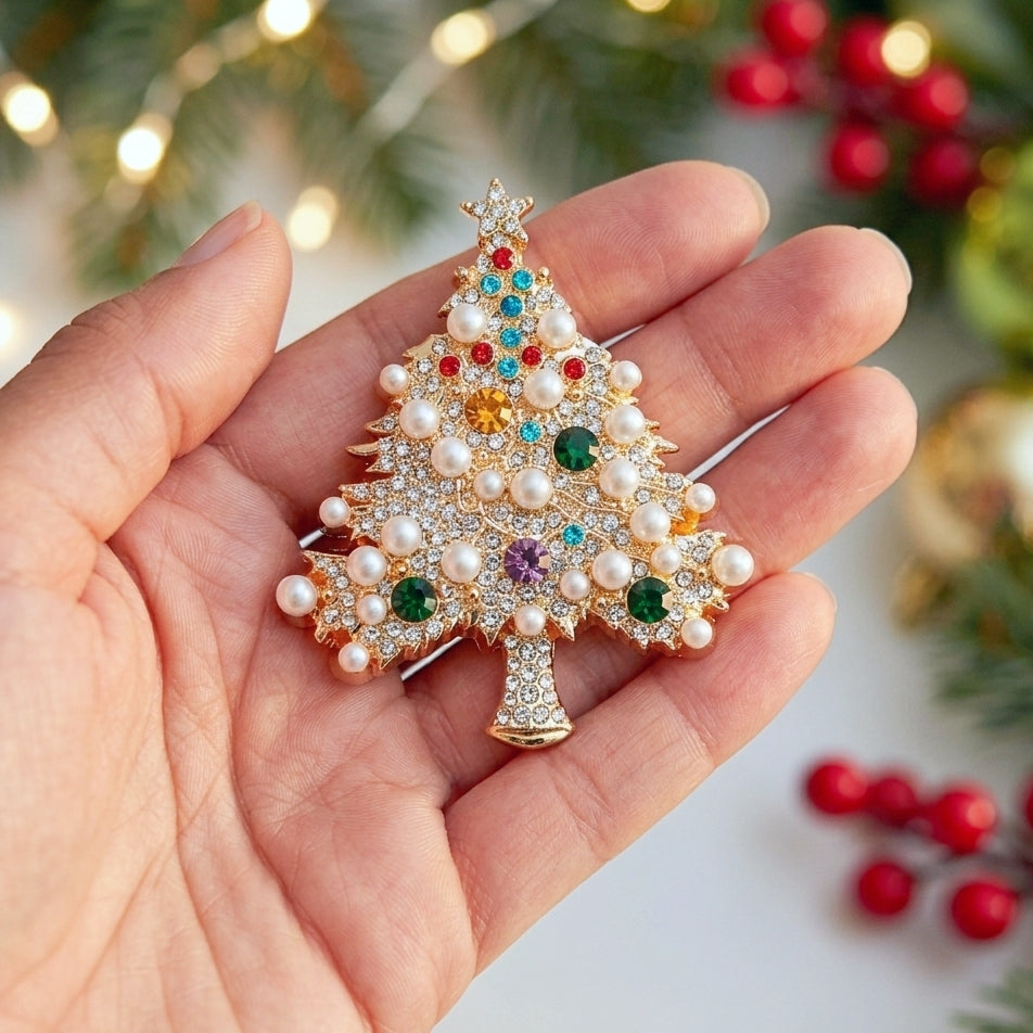 A gold Christmas tree-shaped brooch decorated with white faux pearls and colorful rhinestones, held in a person's hand against a festive blurred background of lights and greenery.