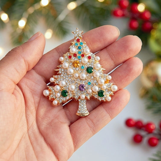 A gold Christmas tree-shaped brooch decorated with white faux pearls and colorful rhinestones, held in a person's hand against a festive blurred background of lights and greenery.