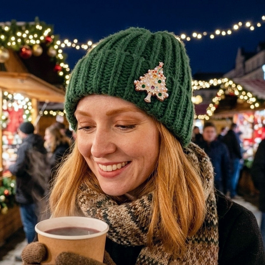 A woman at a Christmas market wearing a dark green ribbed knit beanie with a colorful and sparkly Christmas tree-shaped brooch, a patterned knit scarf, and holding a paper cup of hot gluhwein.