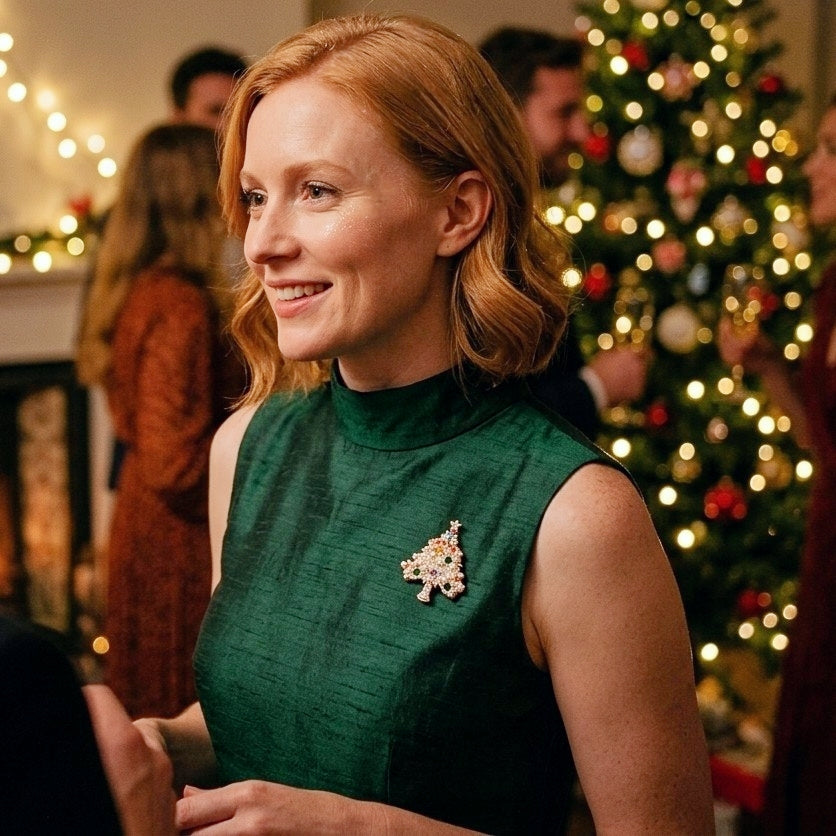 A woman with red hair wearing a dark green sleeveless mock-neck dress and a Christmas tree-shaped brooch, standing in a festive indoor holiday setting with a decorated Christmas tree in the background.