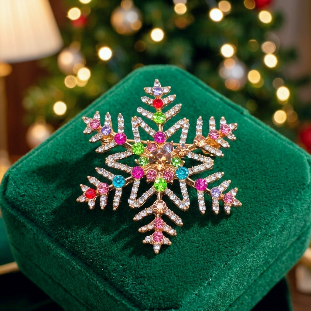 A gold snowflake brooch decorated with pink, green, and blue rhinestones, displayed on a dark green velvet jewelry box with a blurred Christmas tree in the background.