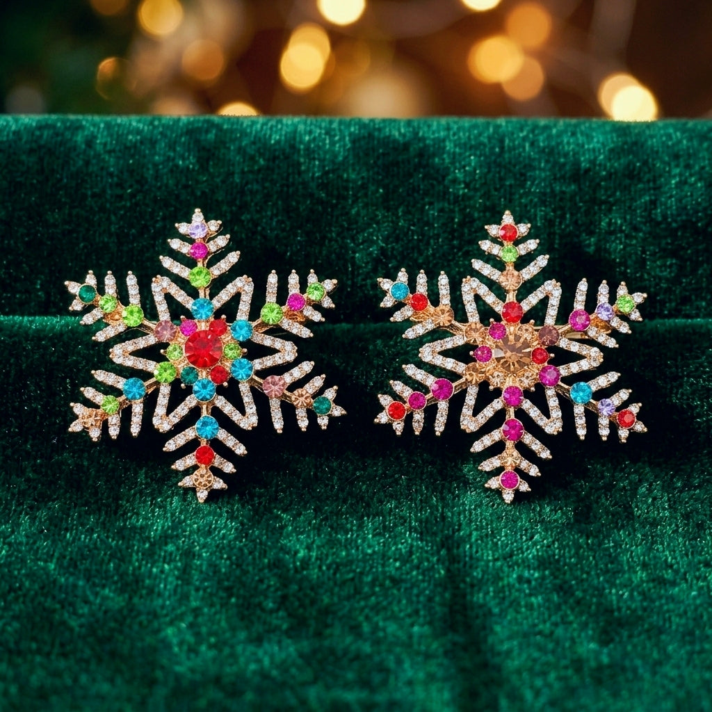 A pair of gold snowflake-shaped brooches decorated with different colorful gemstones, displayed on a dark green velvet surface with a warm, blurred holiday bokeh background.