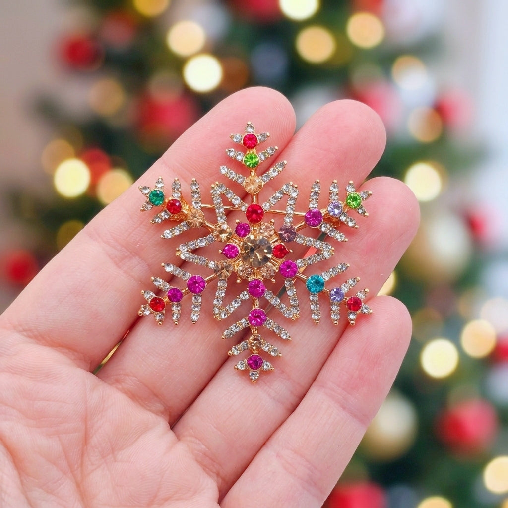 A hand holding a gold snowflake-shaped Christmas ornament decorated with colorful rhinestones and small star accents, with a blurred Christmas tree in the background.