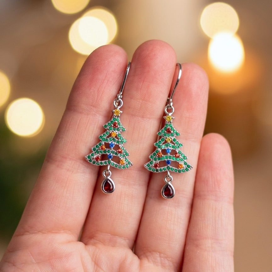 A pair of silver Christmas tree drop earrings with colorful rhinestones and teardrop charms, held between fingers against a warm, blurred bokeh background.