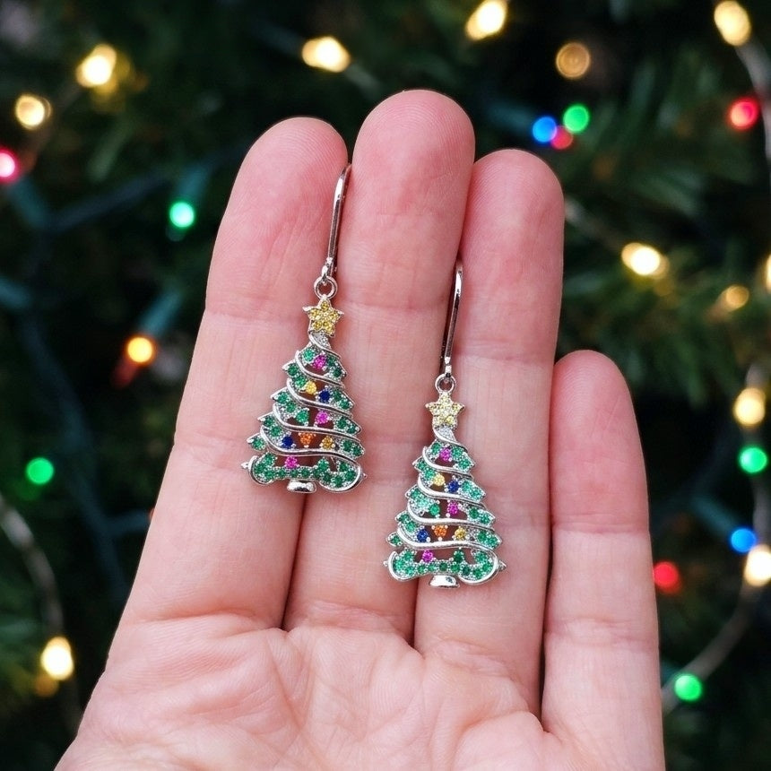 A pair of silver Christmas tree drop earrings with green and multicolor rhinestones, held in a hand against a blurred Christmas tree with colorful lights.