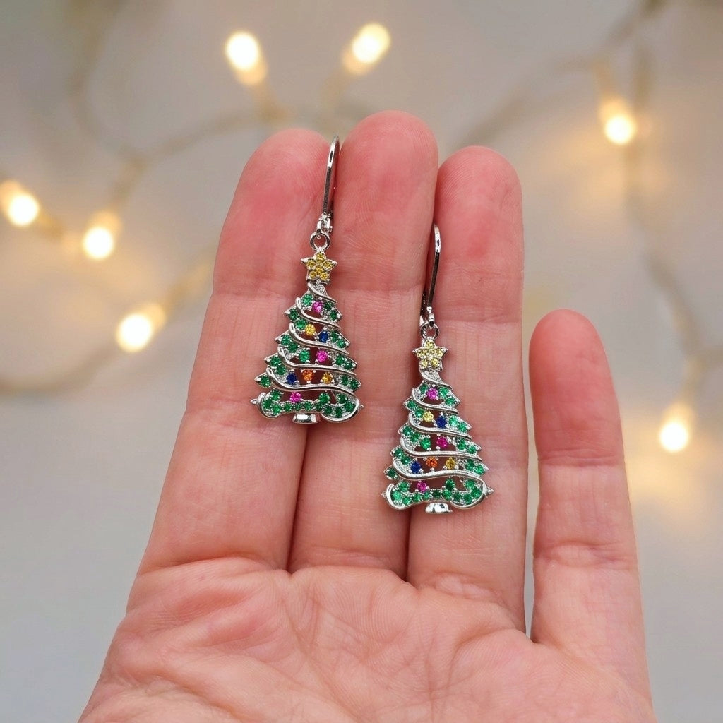 A pair of silver tree-shaped drop earrings with multicolored rhinestones, held between a person's fingers against a soft-focus background with warm string lights.