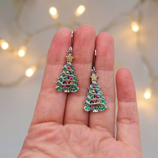 A pair of silver tree-shaped drop earrings with multicolored rhinestones, held between a person's fingers against a soft-focus background with warm string lights.