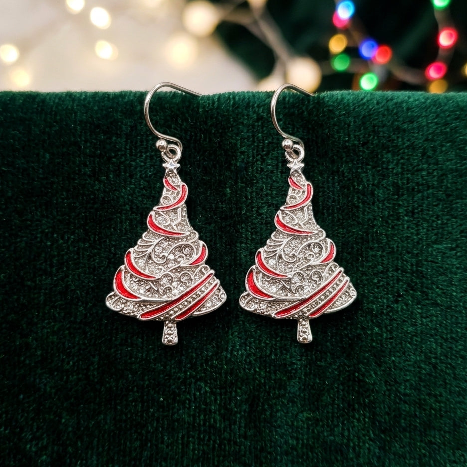A pair of silver Christmas tree-shaped dangle earrings with red enamel stripes, displayed on a dark green velvet surface with blurred holiday lights in the background.