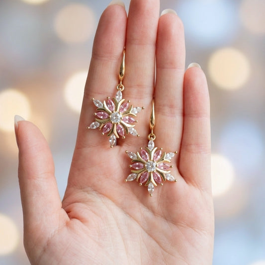 A pair of gold snowflake drop earrings with pink gemstone petals and clear crystal accents, held in a person's hand against a soft, blurred bokeh background.