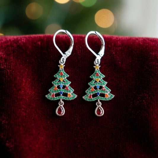 A pair of silver leverback earrings shaped like Christmas trees, decorated with green rhinestones and small red, blue, and orange accents, resting on a dark red velvet surface with a blurred festive background.